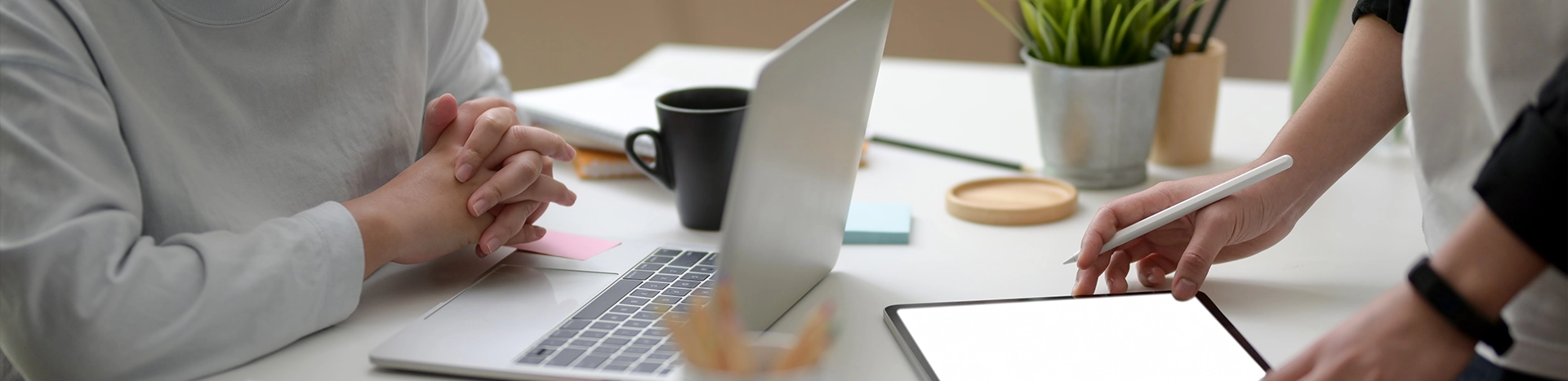 imagen tomada desde un plano bajo, se aprecian manos de personas trabajando sobre una mesa de oficina, con un portatil y una tablet

