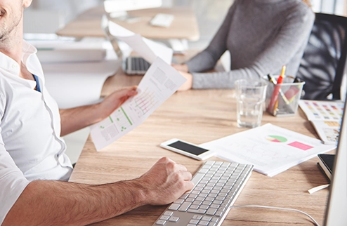 imagen tomada desde un plano bajo donde aparecen cuerpos de personas en oficina, junto a informes con gráficas de colores.
