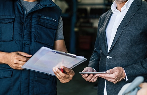 imagen cortada donde se ve el tronco de dos personas negociando, una sujeta un informe con sus manos y lleva chaleco, la otra persona va en chaqueta y sujeta una tablet con sus manos
